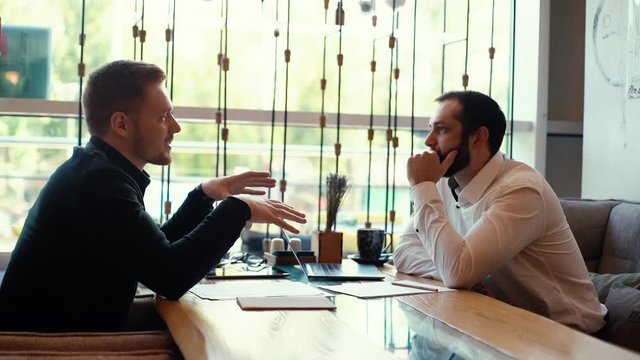 Young business man is talking with client at the table in the interior of the cafe, sitting next to a window. Shot in slow motion
