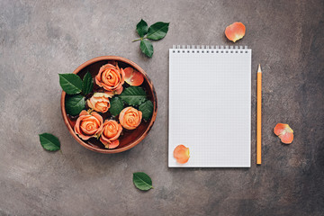 Female workspace with open notepad, ceramic bowl with flowers of coral roses and pencil. Top view, flat lay feminine background.