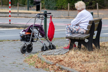 Einsame alte Frau und Großmutter sitzt auf einer Bank am Straßenrand und wartet allein auf den Bus oder ein Taxi mit ihrem Rollator, um die Einkäufe in roter Tasche ins Seniorenheim zu bringen