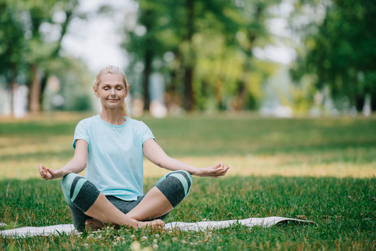 beautiful mature woman meditating with closed eyes while sitting in lotus pose on lawn in park