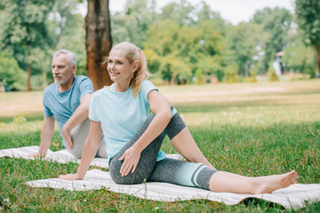 smiling mature man and woman practicing yoga while sitting on yoga mats in park