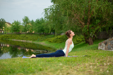 Slim young brunette yogi performs no complicated yoga exercises on the green grass in summer...