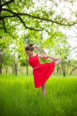 Naklejka na ściany i meble Slim beautiful brunette girl in a red dress performs yoga poses in a summer park. Green forest at sunset