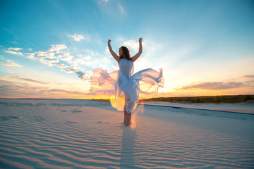 Fototapeta premium A girl in a fly white dress dances and poses in the sand desert at sunset