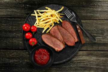 Steak with French fries, fresh cherry tomatoes  and sauce on a rustic wooden table. Wholesome classic American lunch on a black plate. Top view, directly above shot.