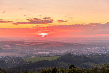 Sunset with view on landscape with fully colored clouds and orange sun goes down and city Valasske Mezirici captured during summer late time