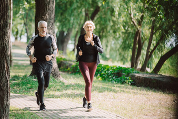 mature, smiling joggers running together on pavement in sunny park