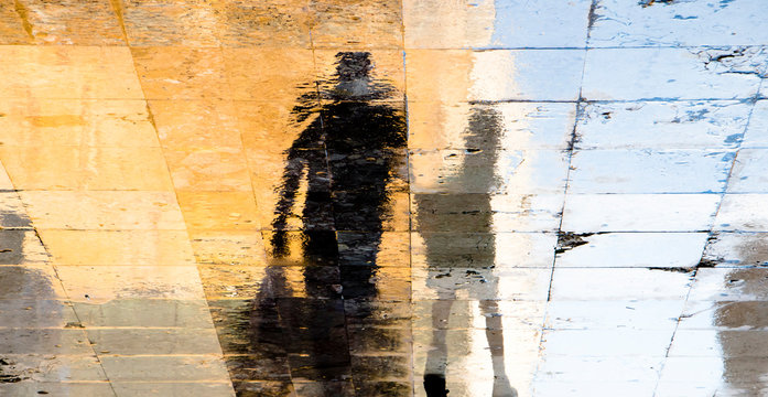 Blurry reflection shadow silhouettes of  a man and a boy walking on a wet street  on a sunny summer day