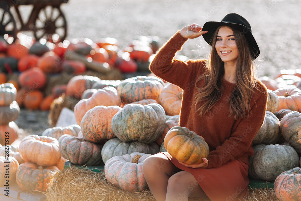 Wall mural Portrait of happy woman sitting on straw on farmers market with orange pumpkin in brown sweater, dress and hat. Cozy autumn vibes Halloween, Thanksgiving day.