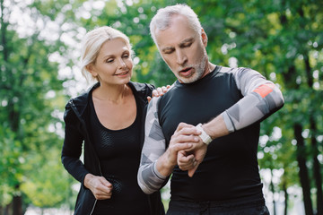 surprised mature sportsman looking at fitness tracker near smiling sportswoman