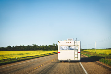The backside of an older pull type camper moving down a highway between canola fields in a countryside summer landscape