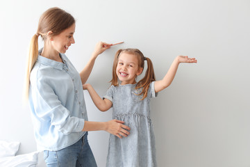 Young mother measuring height of her little daughter near light wall