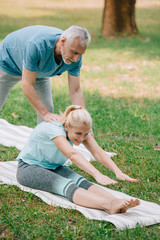 handsome mature man helping smiling woman practicing stretching yoga pose on lawn