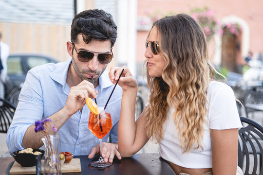 Young couple wearing sunglasses drink with the straw in the same glass a spritz cocktail sitting in an outdoors restaurant. Young people having leisure time drinking alcoholic drinks concept