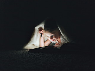 little girl is reading a book under a blanket with a flashlight in a dark room at night