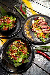 Asian dishes on a black table. Duck breast with peach, noodles with beef, pad thai with chicken and shrimps.
