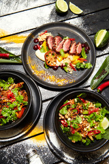 Asian dishes on a black table. Duck breast with peach, noodles with beef, pad thai with chicken and shrimps.