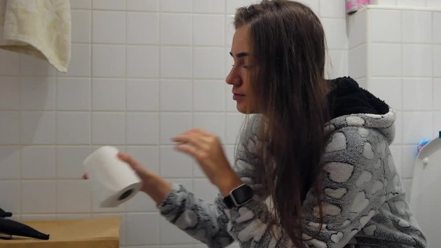 Young woman sitting on toilet in the bathroom and taking toilet paper in hands.