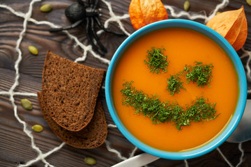 Autumn cream soup garnished with dill in the form of funny faces Halloween pumpkins. Healthy pumpkin and carrot soup puree in a blue bowl on a wooden background. Halloween holiday concept, top view.