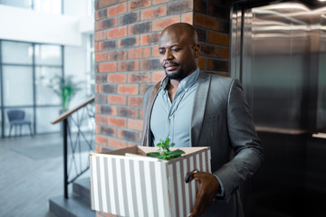 Bearded dark-skinned man leaving office holding box with plant inside