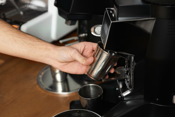 Barista with metal cup using coffee grinding machine in cafe, closeup