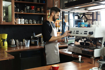 Barista frothing milk in metal pitcher with coffee machine steam wand at bar counter