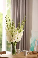 Vase with beautiful white gladiolus flowers, pictures and cups on wooden table in room, space for text