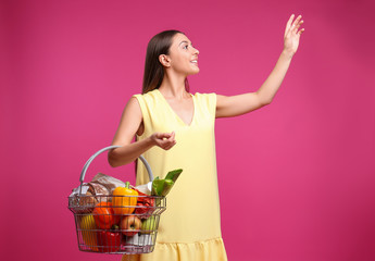 Fototapeta na wymiar Young woman with shopping basket full of products on pink background
