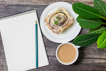 Tasty breakfast in office with traditional Scandinavian dessert cinnabon, cup of coffee and blank notepad on rustic wooden table. Lagom concept.