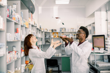 Two funny smiling young pharmacists having fun in pharmacy, throw up different pills