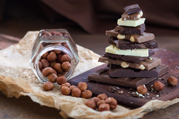 Different grades of chocolate are put the hill on a wooden board, with a filbert in a glass jar on crumpled paper, on a rusty metal surface.