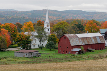 A small church sits on a farm next to a weathered red barn during Autumn in Vermont