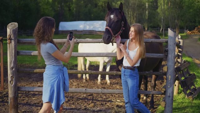 A young woman photographs her friend near a horse in a stable on a summer evening