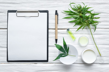 Cannabis cosmetic cream in a jar and a blank paper page with a copy space and a green hemp leaf on a white wooden table background.