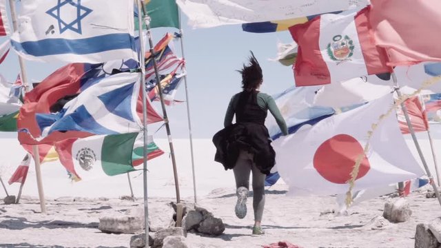 Hispanic woman runs through the countries flags waving with the wind on Uyuni salt flats. Slow motion