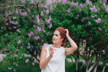 Fototapeta premium Charming woman wearing beautiful white dress standing next to lilac bush. Copy space.