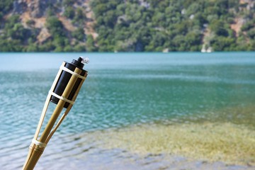 Closeup tiki torch at lakeside, blurred background the surface of the lake with algae and a hill 