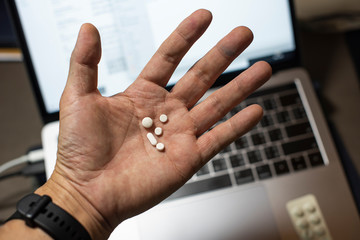 Sick male home office worker taking pills in front of his working space, during his working deadline night. Business or study with stress, late or overwork concepts. Young entrepreneur or student.