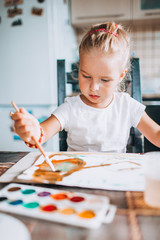 Fototapeta premium Beautiful blonde girl painting with paintbrush and water colors in the kitchen. Kid activities concept. Close up. Toned.