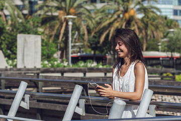 Close-up portrait of cheerful young woman listening to the music on the phone. Beautiful girl over summer background with palm trees in the city. Gorgeous brunette lady enjoying the music outdoors.
