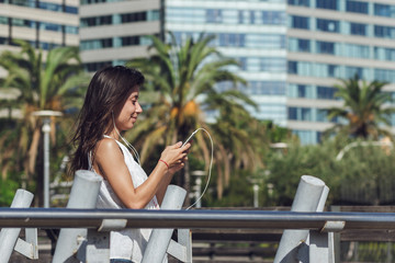 Fototapeta premium Portrait of beautiful girl listening to the music and reading a message on her smartphone outdoors. Close-up photo of gorgeous brunette young woman over summer background with palm trees and buildings