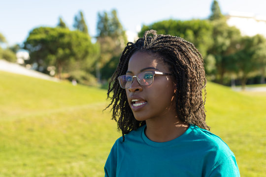 Excited Volunteer Working Outside. Young Black Woman Wearing Uniform And Eyeglasses Standing On City Lawn, Looking Away And Speaking. African American Volunteer Concept