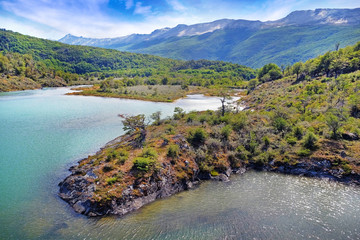 Panoramic view of Tierra del Fuego National Park, showing mountains surrounded by green vegetation and water, against a blue sky.