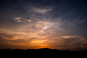Twilight dramatic cloudy blue sky with orange sunlight and silhouette of mountains.