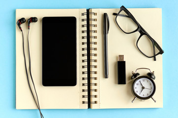 Fototapeta premium Black glasses, cellphone, paper notepad, alarm clock and headphones on blue background composition.
