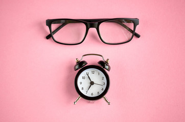 Black glasses and alarm clock on pink background composition.