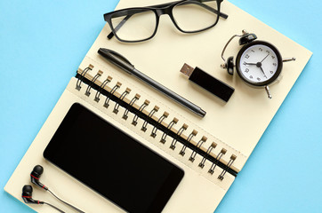 Black glasses, cellphone, paper notepad, alarm clock and headphones on blue background composition.