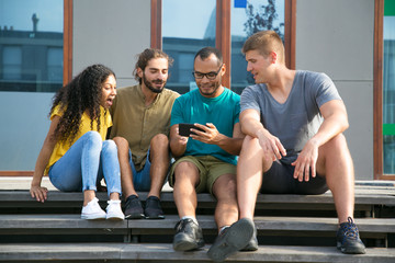 Excited friends watching video on mobile phone. Cheerful Latin guy using smartphone and showing screen to young man and woman outside. Watching content on phone concept