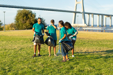 Team of cleaning workers removing trash from grass. Men and women wearing uniforms working with rakes and holding plastic bags with trash. Outdoor cleaning concept