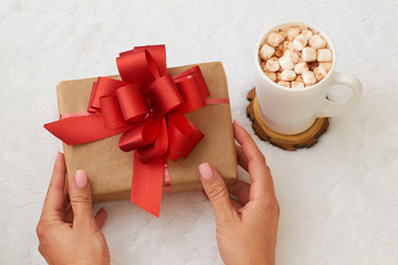 A woman holds a gift with a red bow in her hands, next is a cup of fragrant cocoa with marshmallows